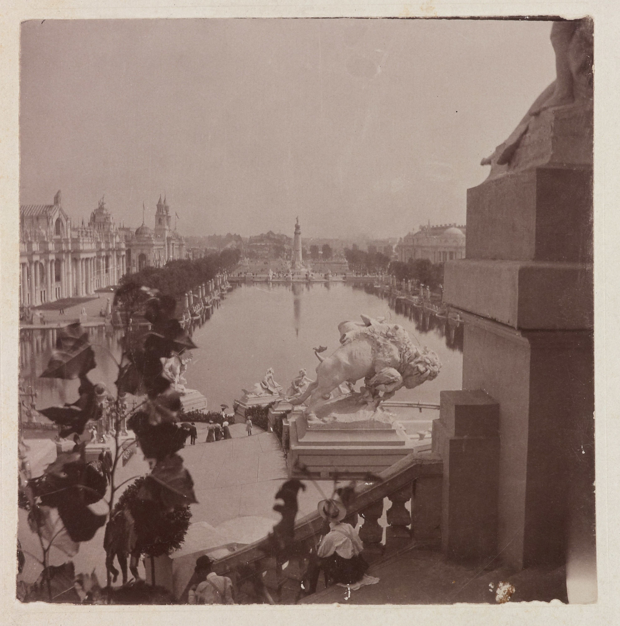 A black-and-white photograph of a tree-lined reflecting pool taken from the steps of a building showing other buildings and several sculptures, including a bison, lining the pool.