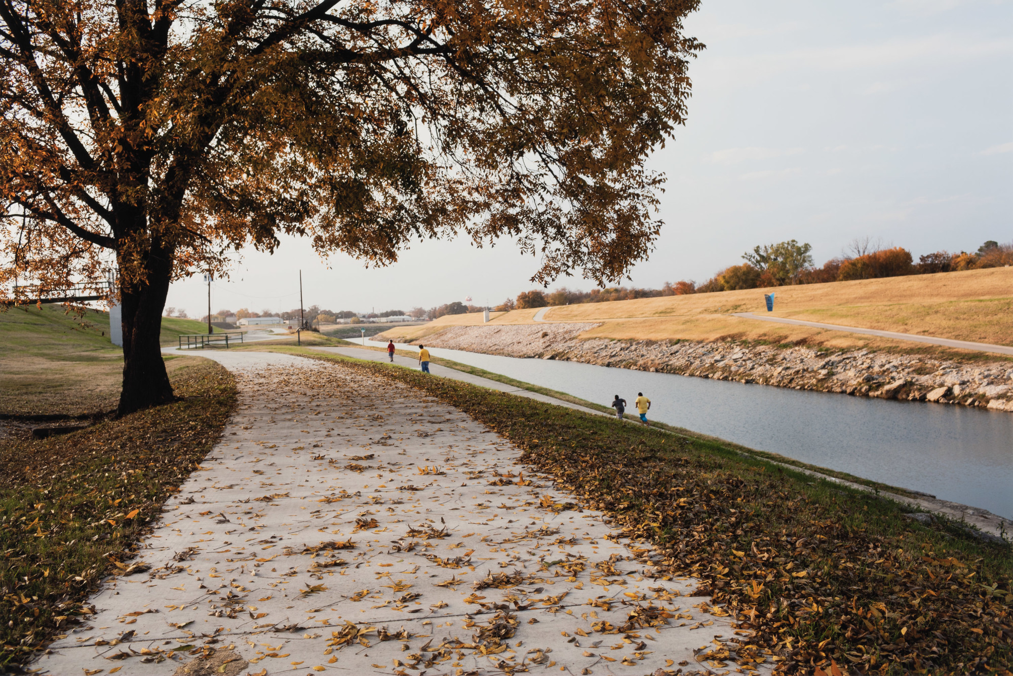A color photograph of a late fall landscape with a river disappearing into the distance and people running on well-groomed paths.