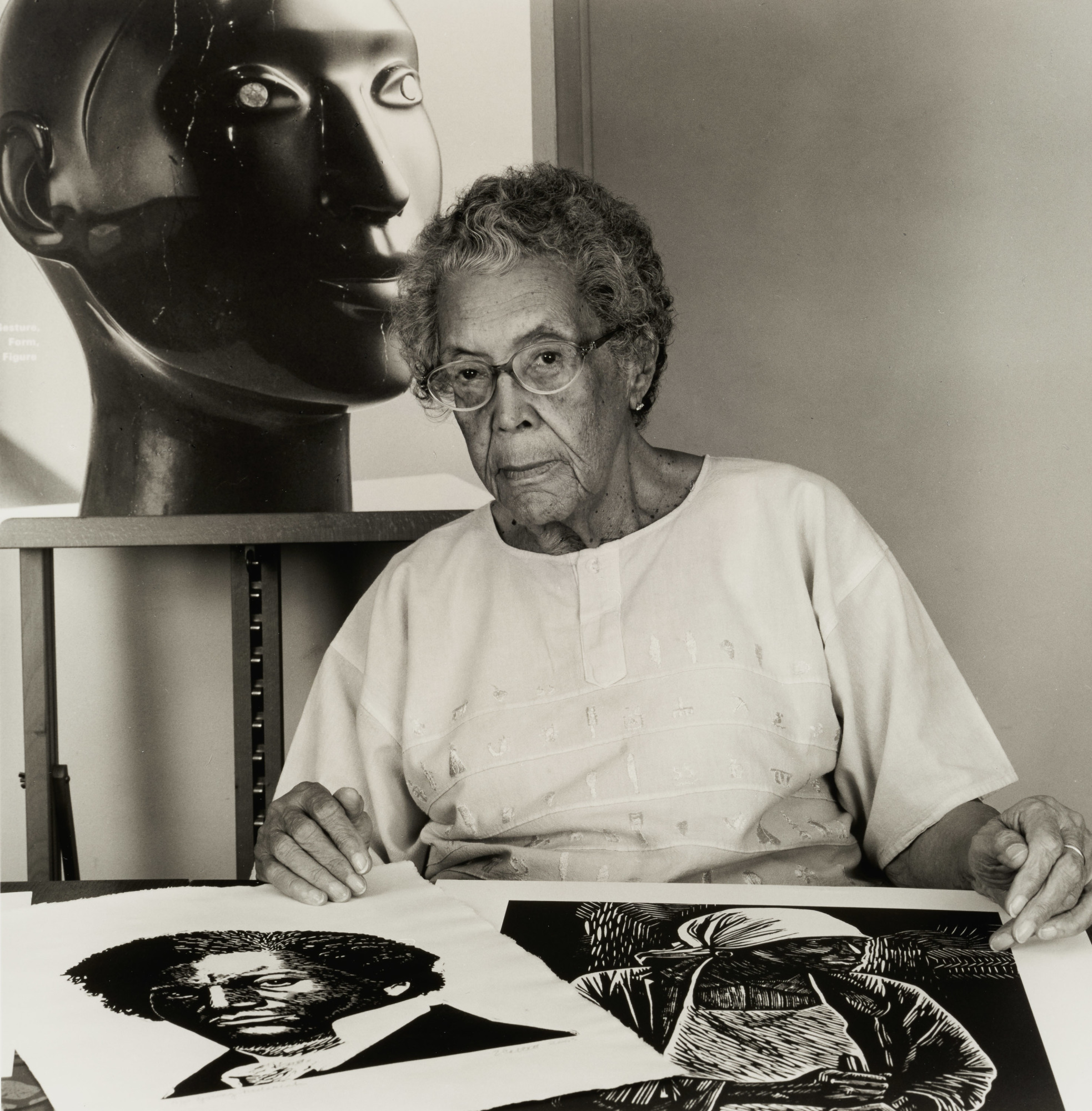 A black-and-white photograph of an older Black woman with two prints on a table in front of her and a large black sculpture of a head behind her.