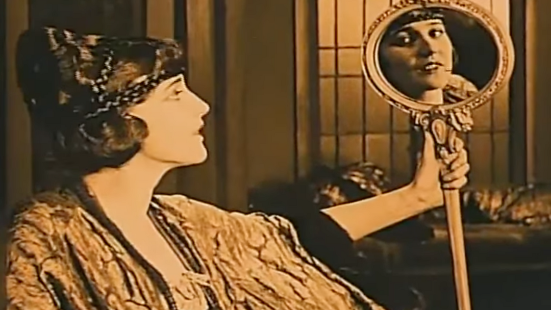 A screenshot from a movie of a White woman holding a mirror that shows her reflection.
