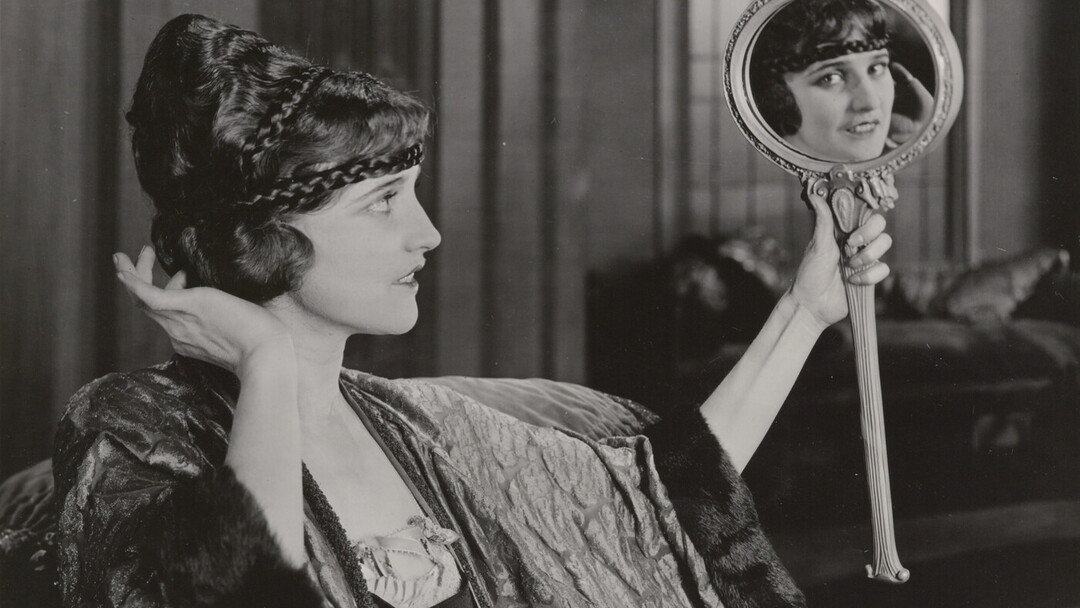 A black-and-white photograph of an elaborately dressed White woman holding up a mirror and gazing into it, her gaze in the mirror meeting the viewer&#039;s eye.