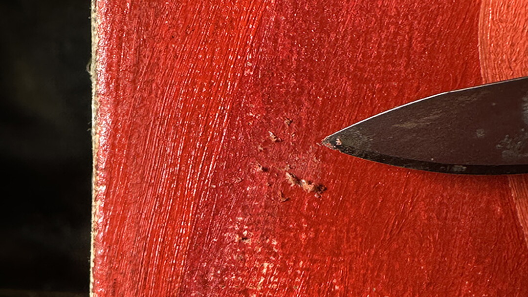 A close-up of a red painting with a scalpel pointing to streaks in the paint layers.