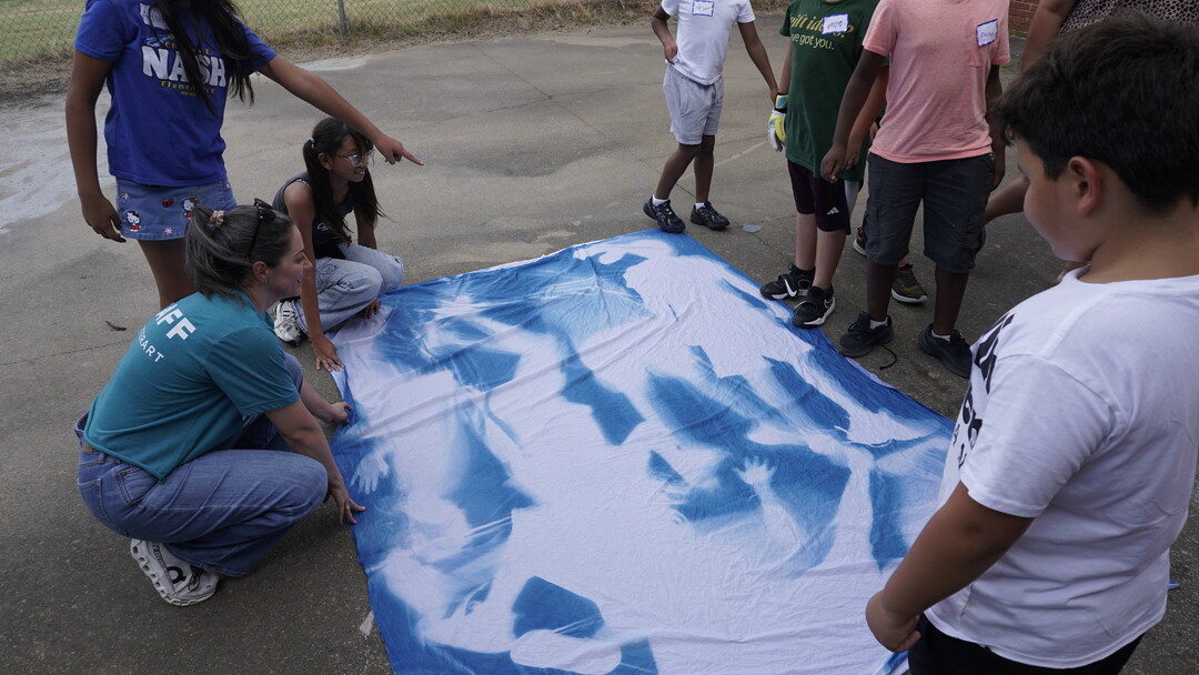 A group of middle-school-aged children and an adult lay out a large piece of fabric with blue silhouettes.