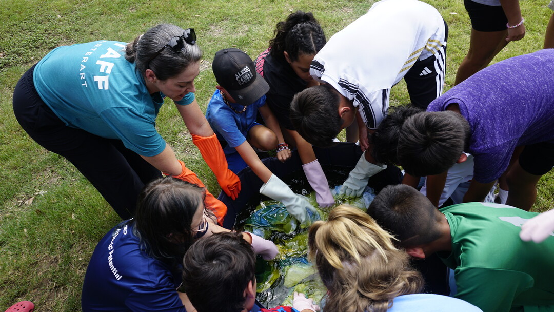 A group of middle-school-aged children, all wearing rubber gloves, rinse paper in a tub of water.