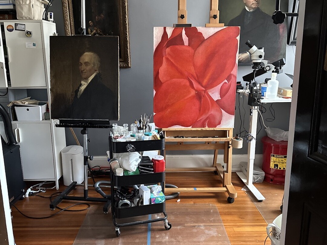 A painting of a red flower sits on an easel in a studio filled with other paintings.