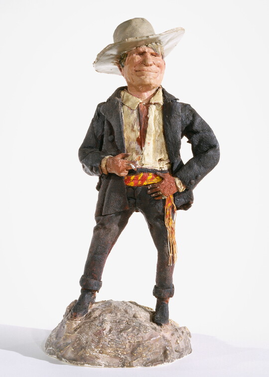 A painted sculpture of a White man wearing a cowboy hat, a light-colored shirt, and dusty pants and jacket standing on a rock, smoking a cigarette.