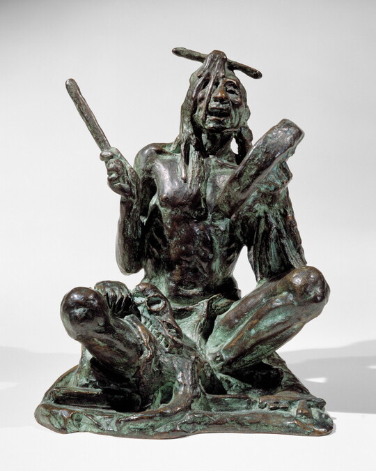 A bronze sculpture of a sitting Indigenous man playing a handheld drum.