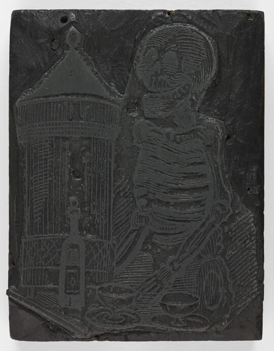 An engraved wood printing block depicting a skeleton standing in front of a table with a bottle and two glasses.