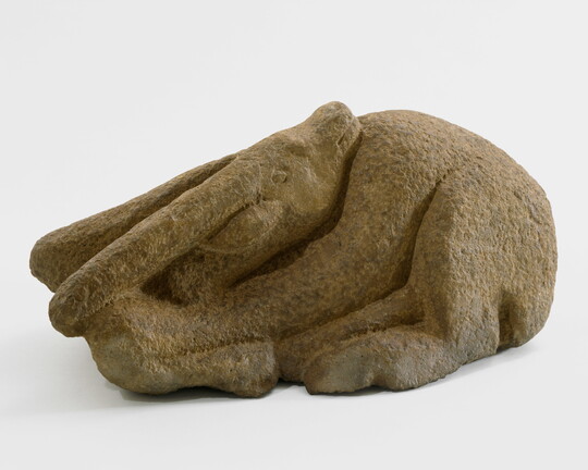 A stone sculpture of a goat curled on the ground with its head turned so its chin rests on its shoulder.