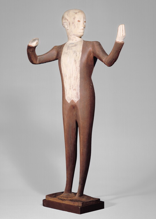 A wood sculpture of a male orchestra conductor standing with his arms raised out to his side.