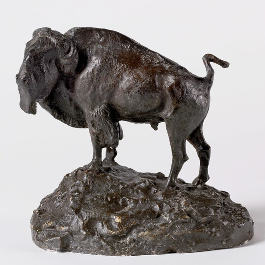 A brown sculpture of a bison standing on a slightly rocky incline.