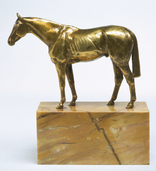 A yellow bronze sculpture of a muscular horse with a long tail on a base of yellow marble. 
