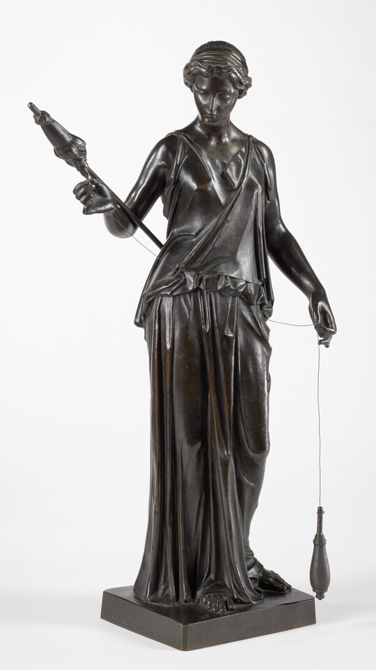 A bronze sculpture of a woman holding a staff in one hand and spooling thread onto a spindle with the other.