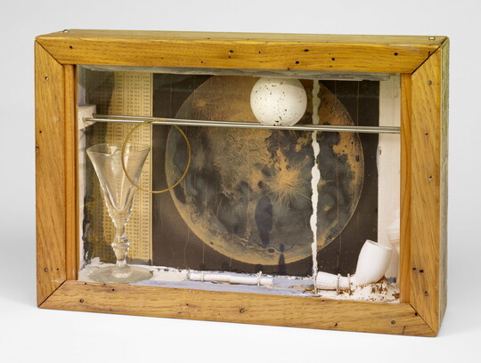 A wood shadowbox containing a pipe, a white ball with gray speckles, a picture of the moon, and a glass goblet.