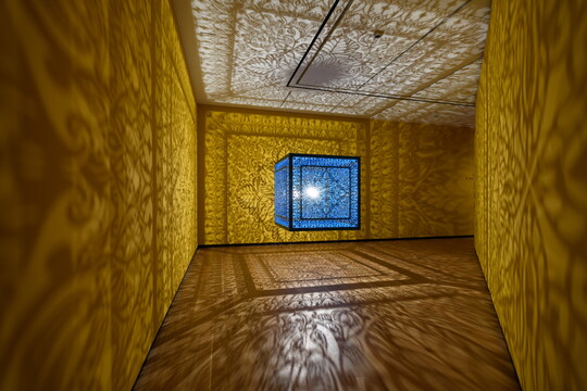 A brightly lit and suspended blue cube with laser cut shapes on all sides that cast intricate shadows on yellow walls.