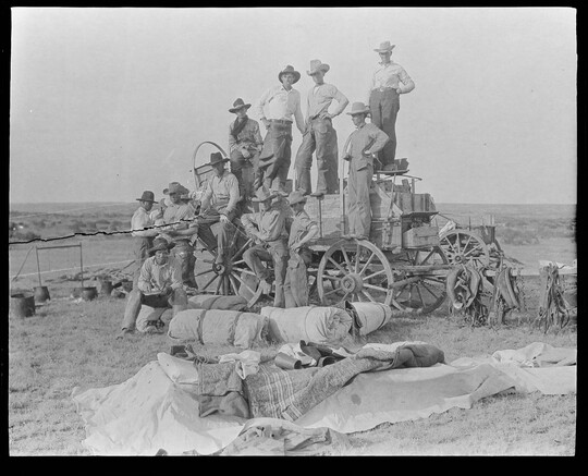 A black-and-white image of a group of cowboys posed on a wagon, bedrolls and blankets on the ground in front of them.