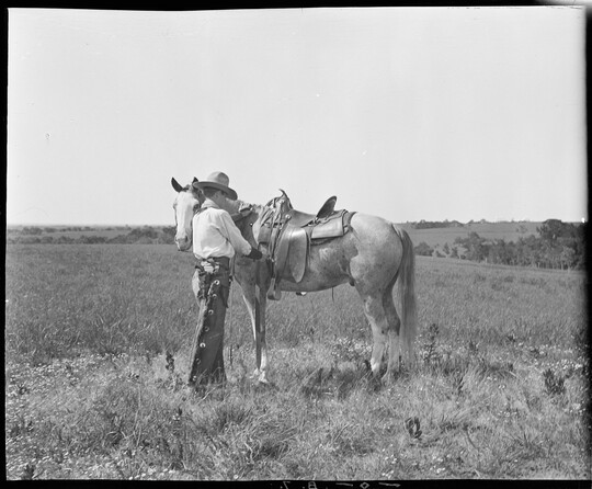 A black-and-white image of a cowboy standing in a field next to his horse adjusting the saddle.