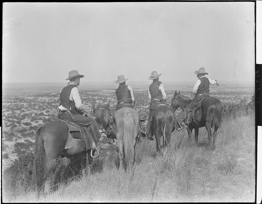 A black-and-white photograph of four cowboys on horseback, viewed from behind, looking over plains in the distance.