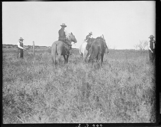 A black-and-white image of a couple of cowboys on horseback and two standing in a grassy field.