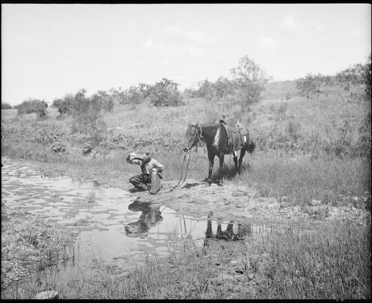 A black-and-white image of a cowboy crouched next to a stream drinking water from his hat as his horse stands behind him.