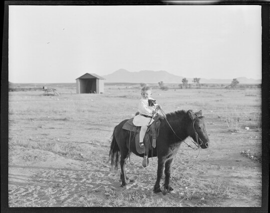 A black-and-white image of a young White child on a pony in a dusty field.