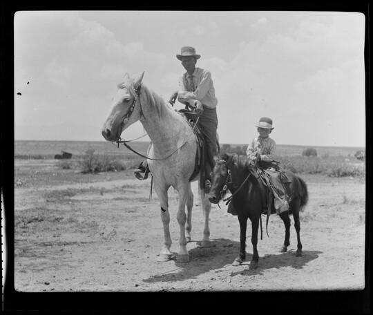 A black-and-white image of a White man wearing a cowboy hat on horseback and, next to him, a White child wearing a cowboy hat on a pony.