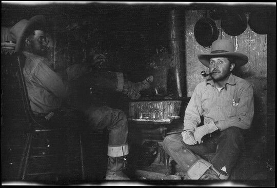 A black-and-white image of two White cowboys seated inside: one on a chair with his foot propped on a stove, the other next to the stove smoking a pipe.