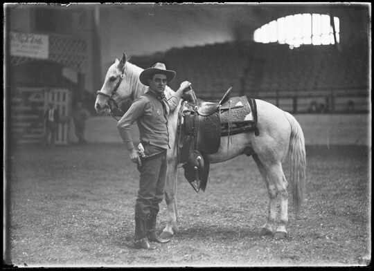 A black-and-white image of a White cowboy standing next to his saddled horse in an empty arena.