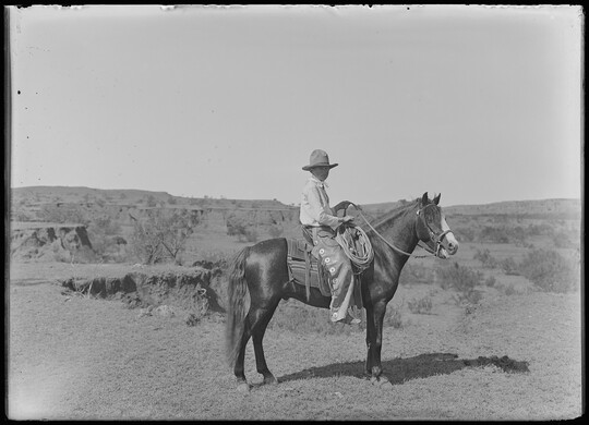 A black-and-white image of a cowboy on horseback standing, in profile, on a plain.