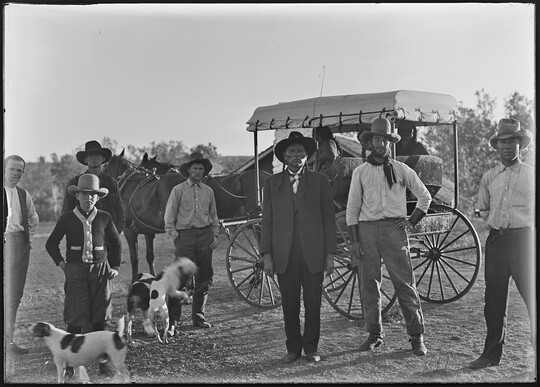 A black-and-white image of six men dressed in western attire and several dogs standing in front of a carriage.