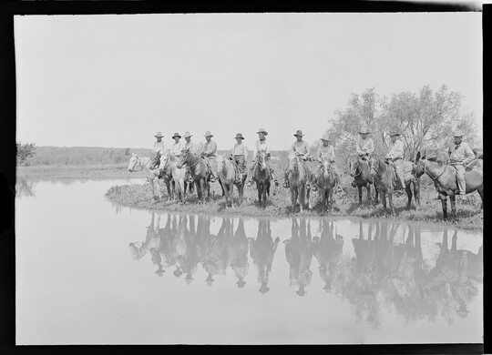 A black-and-white image of a group of cowboys on horseback lined up along the shore of water.