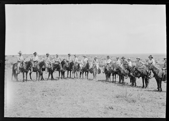 A black-and-white image of a group of cowboys on horseback lined up on a grassy plain.