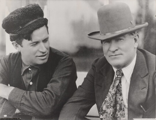 A black-and-white photograph of two White men: the man on the left wearing a fuzzy hat, dark vest, and button-down shirt; the man on the right wearing a cowboy hat with a jacket and tie.