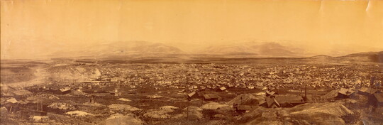 A sepia-toned panoramic photograph taken from high up of a small town in a barren, rocky landscape with mountains in the distance.
