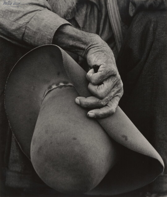 A close-up black-and-white photograph of a hand with aged, wrinkled skin holding a felt cowboy hat.