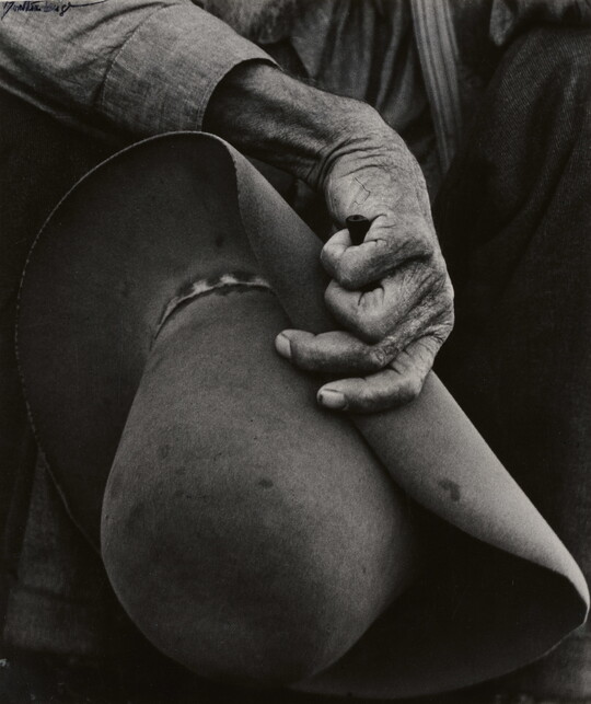 A close-up black-and-white photograph of a hand with aged, wrinkled skin holding a felt cowboy hat.