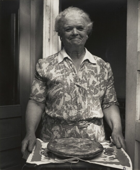 A black-and-white photograph of a smiling older White woman wearing a floral print dress and holding out a tray with a pie on it.