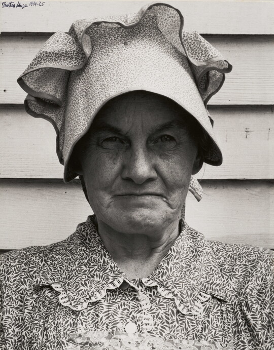 A black-and-white photograph of an older White woman, head and shoulders filling the frame, wearing a bonnet and a printed blouse.
