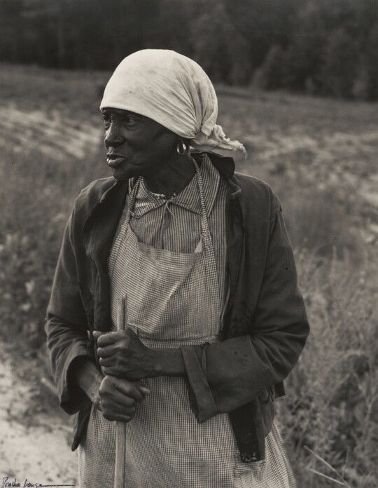 A black-and-white photograph of a Black woman wearing frayed clothing and a white head scarf holding a wood rod in a field.