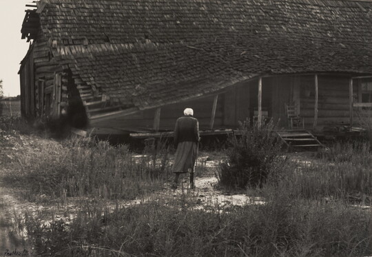 A black-and-white photograph of a hunched figure walking toward a log cabin with a collapsed roof and a rocking chair on the porch.
