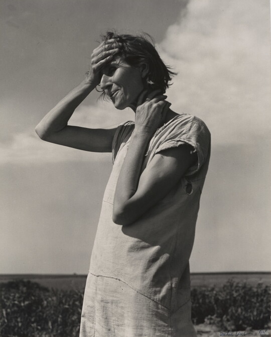 A black-and-white photograph of a White woman standing outside, holding the side of her neck with one hand and her forehead with her other.