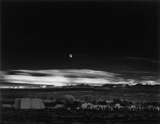 A black-and-white landscape photograph of a town with mountains in the background and the moon rising above the clouds in the night sky.