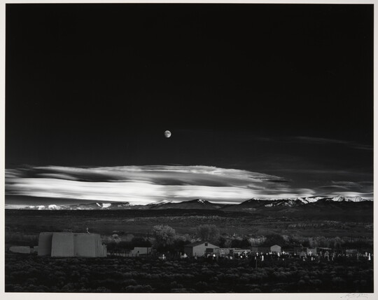 A black-and-white landscape photograph of a town with mountains in the background and the moon rising above the clouds in the night sky.