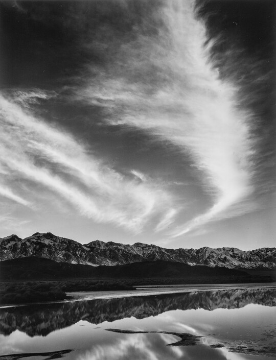 A black-and-white landscape photograph of mountains and clouds that reflect on the still water in the foreground.