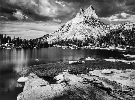 A black-and-white photograph of a rocky mountain peak from across a still lake surrounded by trees.