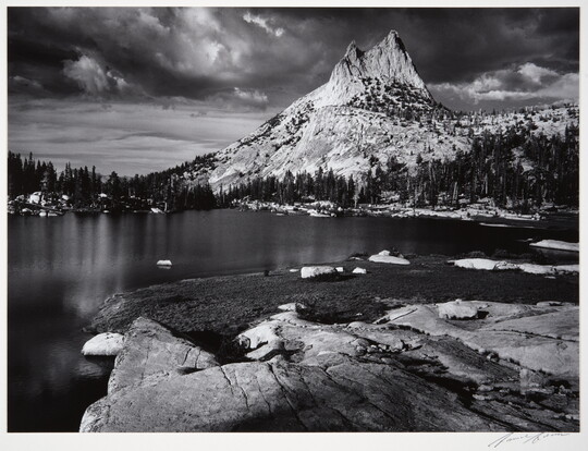 A black-and-white photograph of a rocky mountain peak from across a still lake surrounded by trees.