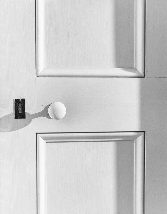 A black-and-white close-up photograph of a white, paneled door, with a metal keyhole on the left side and a white doorknob to the right of it.