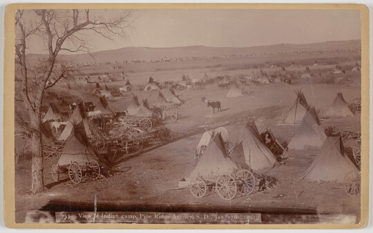 A sepia-toned photograph of many tipis and wagons on a plain.