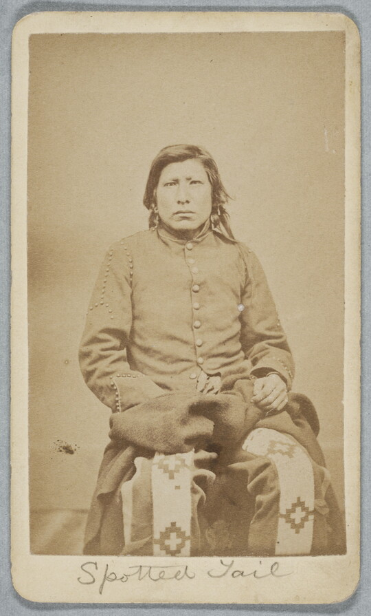 A black-and-white photograph of a seated Indigenous person with mid-length dark hair, wearing a long sleeved coat with large buttons and wide patterned pants.