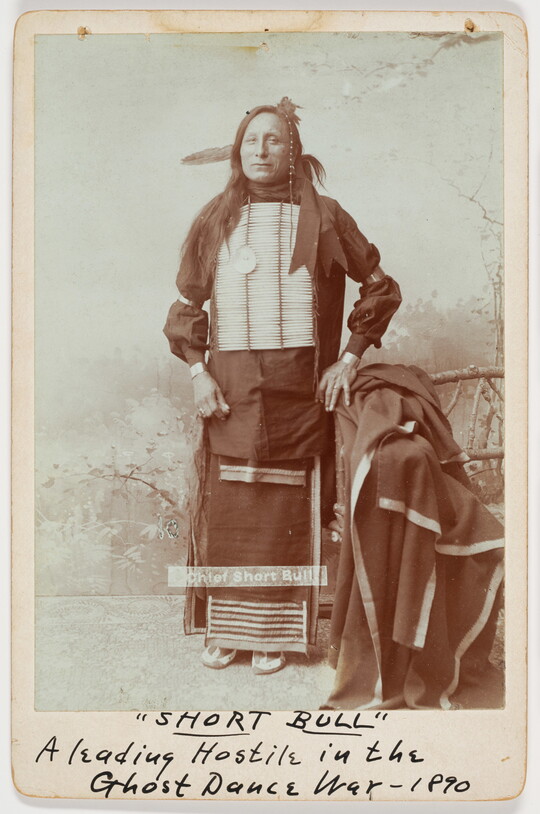 A sepia-toned full-body portrait of an Indigenous man in cultural dress, including a breastplate.
